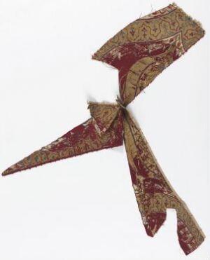  A fragment of an antique fabric in faded burgundy with intricate mustard yellow patterns, displaying signs of aging with frayed edges and loose threads.