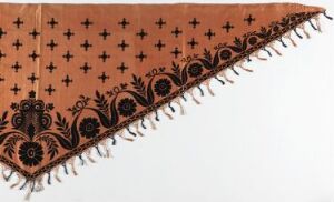  Part of a terracotta or rust-colored textile decorated with a pattern of dark brown crosses and a detailed paisley border with light-colored fringes on the edge. The fringes hang freely, offering a textural contrast to the warm, earthy color of the fabric.