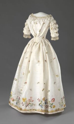  An historical cream-colored dress with a fitted bodice and full skirt, adorned with intricate multi-colored floral embroidery, displayed against a gray background.