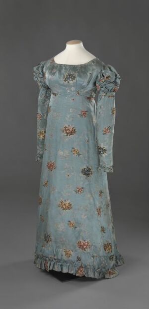  An early 20th-century pastel blue vintage dress with a floral pattern and lace trim displayed on a mannequin against a grey background. Artist name and title are unknown.