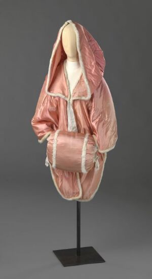  A pale pink, hooded, silk-like puffed garment with a subtle sheen is displayed on a featureless mannequin against a dark gray background. The piece is elegant, draping luxuriously with a cinched bubble hem and wide, elbow-length sleeves.