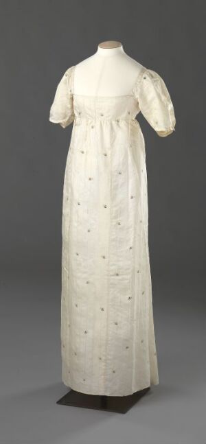  An early 19th-century Regency style dress with a high empire waist and short puffed sleeves, in an off-white or cream color with a subtle floral pattern, displayed on a featureless mannequin against a gray background.