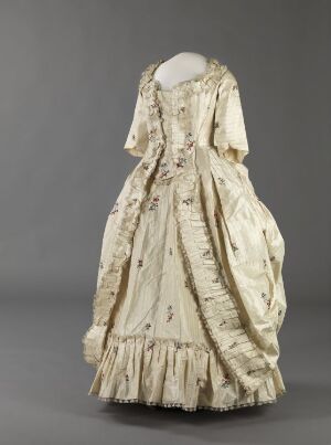  A historical 18th-century off-white dress with a square neckline and elbow-length sleeves, adorned with vertical stripes and floral patterns, ruched swags on the skirt, and a ruffled hem, displayed against a gray background.