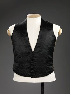  A classic black men's vest on an ivory mannequin torso against a gray background. The vest has a deep V-neck with a buttoned front and a smooth, shiny texture, suggestive of a formal wear garment.