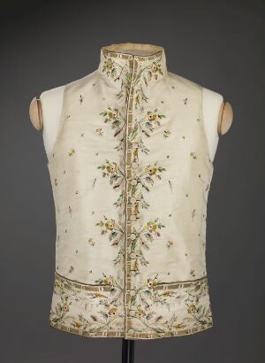  A cream-colored, sleeveless waistcoat with detailed green and gold floral embroidery displayed on a torso mannequin, set against a dark grey background. The vest features an ornate design with a high collar and an embroidered horizontal band around the waist.