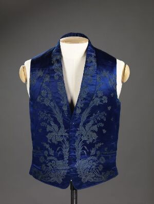  A royal blue gentleman's vest with a dark blue floral and acanthus leaf pattern on a torso mannequin against a grey background. The vest has no visible fastenings and features a V-neck cut. Artist name and title are unknown.