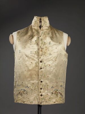  An antique cream-colored vest with faded golden embroidery, featuring a high collar and front button closure, displayed on a mannequin against a gray background.