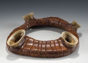  A wooden jewelry piece by artist Liv Blåvarp, featuring a curved, segmented design with rich mahogany segments transitioning into lighter holly wood at the ends. The woodworking technique is detailed, suggesting a polished and finely crafted object, possibly intended as a statement bracelet or neckpiece.