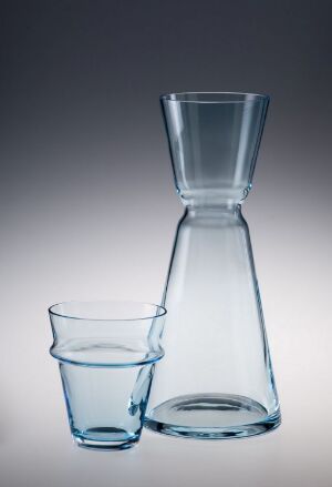  A carafe and tumbler made of blown colored glass with a light blue tint, showcasing a minimalist design typical of Magnor, Norsk Glassverk A/S. The background has a gradient from light to dark, emphasizing the translucency and color of the glassware.