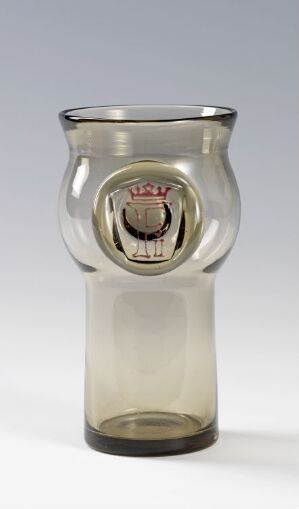  A decorative blown glass object with a tapered cylindrical shape, featuring a central emblem in red, white, and gold colors, likely representing a coat of arms, encased within the clear to smoky gray glass, against a neutral background.