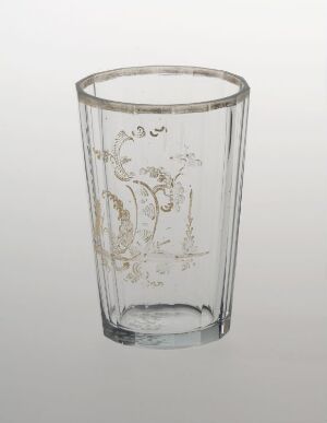  Transparent drinking glass with a tapering cylindrical shape, decorated with delicate gold or light brown floral or vine-like motifs, against a plain light-gray background.