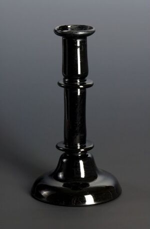  A glossy black candlestick holder with a round base and a tapered stem featuring decorative nodes, set against a neutral gray background. Its reflective surface subtly mirrors its surroundings. Artist name and title are unknown.