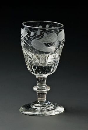  An intricately etched clear crystal wine glass with floral design stands against a dark gray background, reflecting light on its smooth surface. Artist name and title are unknown.