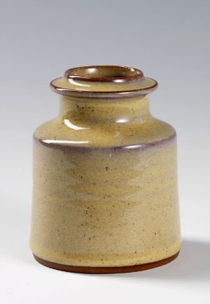  A small, handmade ceramic jar with a simple design, featuring an off-white to honey gradient glaze and sporadic brown speckles, against a plain light gray background.