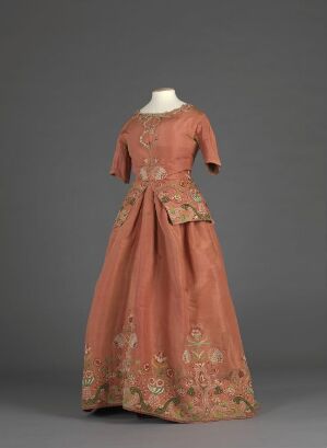  A historic terracotta-colored dress with a floral pattern, fitted bodice, and full skirt, displayed against a gray background.