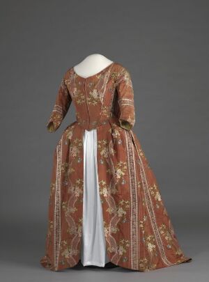  An antique rust-colored dress with an elaborate floral pattern on display, featuring a full skirt, fitted bodice, and contrastingly bright white panel at the front of the skirt, against a neutral gray background. Artist name and title are unknown.