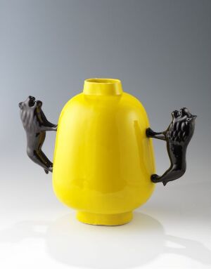  A vivid yellow porcelain vase by artist Heidi Bjørgan with a glossy finish and two stylized, matte black figures serving as handles on its sides.