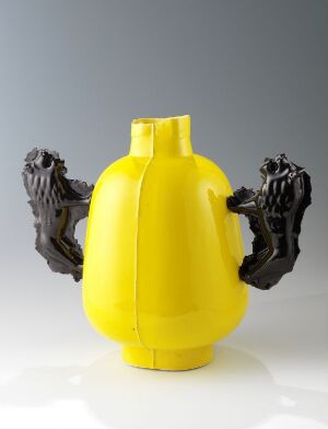  A porcelain artwork by Heidi Bjørgan consisting of a bright yellow segmented vessel with two realistic black animalistic handles attached to its sides, set against a neutral grey background.