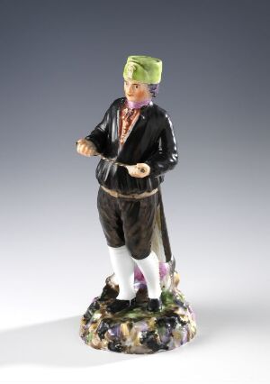  Figurine of a person wearing a green turban, black jacket with golden details, white breeches, pink stockings, and black pointed shoes, standing on a colorful base suggestive of natural foliage.