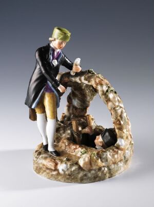  A porcelain figurine showing two characters in a scene, one standing in a royal blue and gold coat offering a handkerchief, with a powdered wig, the other crouching in a rock formation, finished with glossy and detailed coloring.