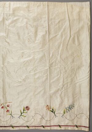  A cream-colored, rectangular fabric with natural creases and a vertical fold line, adorned with a row of delicate, hand-embroidered flowers in reds, yellows, greens, and touches of blue along the bottom edge, finished with a thin wavy red line.