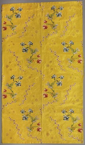  Vertical, rectangular image of a decorative fabric or paper with a mustard yellow background featuring symmetrical patterns of blue, red, and white flowers intertwined with green vines. A central vertical seam or fold runs through the entire length of the piece.