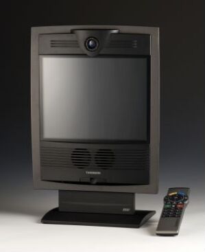 Alt-Text: A modern TV set by Roy Tandberg, with a large, square, dark screen, a speaker grille below and a possible built-in camera above. The TV is placed on a dark pedestal stand and accompanied by a long, slender remote control with white, gray, and red buttons, against a nondescript background.