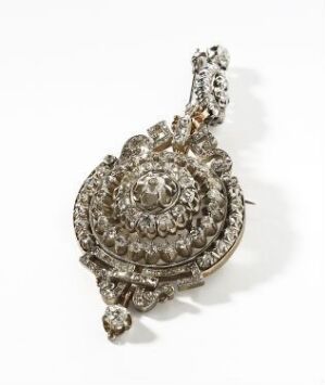  An ornate piece of silver or white gold jewelry, possibly a brooch or pendant, with a central large clear gemstone surrounded by multiple tiers of smaller stones, creating a dazzling concentric pattern on an off-white background.