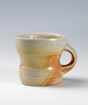  A handcrafted ceramic mug with a gradient glaze that transitions from toasted honey at the bottom to beige and then ivory at the top, featuring a contrasting terracotta glazed handle, set against a light-grey background.