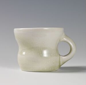  A simple, off-white ceramic mug with a hint of celadon green, featuring a rounded body and a single handle, set against a neutral gray background. Artist name and title are unknown.