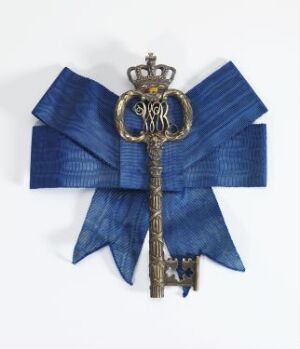  A ceremonial key "Kammerherrenøkkel" made of cast and gilded silver with pierced decoration. The key features a crown atop its bow and is adorned with a broad ribbed blue silk ribbon with a moiré effect. The ornate design and luxurious materials indicate its historical and ceremonial importance, crafted by an unidentified artist.