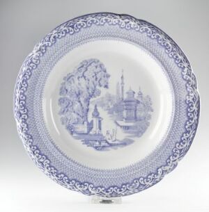  A glazed ceramic plate from Egersunds Fayancefabrik with blue and white traditional design. The plate features a central pastoral scene with a tree, buildings, and a bridge, framed by a decorative floral border.