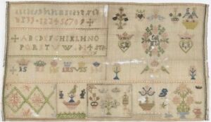  An antique embroidery sampler featuring alphabets at the top, followed by rows of various stitched patterns including flowers, leaves, geometric designs, and coat of arms emblems in a muted color palette on an aged, off-white fabric.