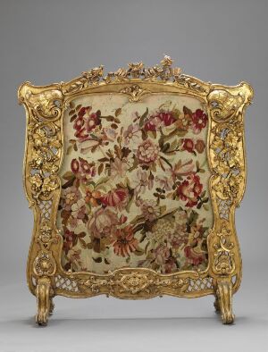  Antique gilded fire screen with a central floral tapestry in red, pink, green, and brown on a creamy beige background, surrounded by an ornate gold-colored frame with baroque or rococo style motifs, standing on scrolled feet.