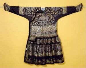  A traditional garment with intricate metallic patterns on a yellow background. The garment has wide sleeves and a T-shaped silhouette, featuring detailed patterns in dark tones accented by symbolic motifs and coin-like elements, hinting at cultural significance and craftsmanship.