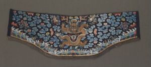  An embroidered textile with a crescent shape, featuring a dense floral pattern in shades of pink and blue on a navy background, with a central emblem in gold tones suggestive of an ornate vase or urn.