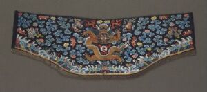  A semi-circular traditional textile piece with a dark blue background featuring dense floral patterns in various shades of blue with pink, white, and red accents. A stylized brown animal is centered in the arch, framed by white wave-like patterns at the lower edge. Artist name and title are unknown.