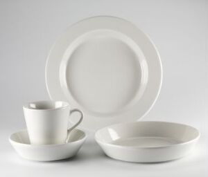  A set of vitreous porcelain dinnerware designed by Ingegerd Råman, including a large white glossy flat plate, a smaller white deep plate, and a white cup with a saucer, all presented against a light grey background.