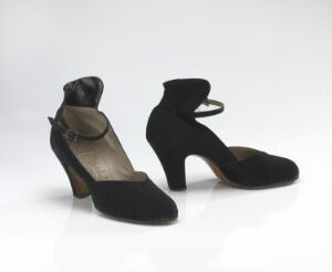 
 A pair of classy black high-heeled shoes with a smooth, velvety texture and a round-toe design, featuring ankle straps and a subtle decorative detail on the front, set against a light gray backdrop.