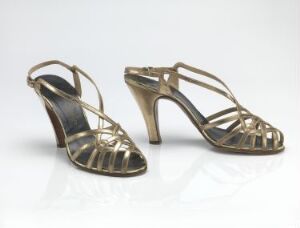  A pair of metallic gold high-heeled shoes with strap design on a reflective light surface against a white background.