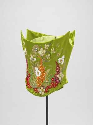  A design piece by Ove Harder Finseth titled "Korsett," featuring a machine-woven, hand-embroidered lime green silk corset, ornately detailed with glass stones, metal and plastic sequins, silk and gold thread embroideries, and plastic boning displayed on a plain black stand against a neutral backdrop.