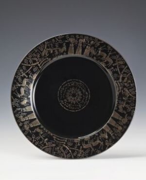  A decorative plate with a dark black background and a silver-gray ornate border, featuring intricate symmetrical patterns with geometrical and vegetal elements. The center of the plate is a solid black, contrasting with the detailed rim.