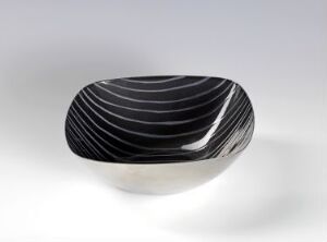  A Grete Prytz Kittelsen-designed stainless steel bowl with pressed enamel black stripes, creating a contrasting and rhythmic design on the reflective metal surface. The bowl's shape widens from a narrow base to an open top, with glossy stripes emphasizing its form.