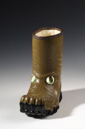  A children's olive green rubber boot designed by Viking Fottøy A/S, resembling a dinosaur foot with textured scales, playful eyes, and a sturdy sole, standing against a grey background.