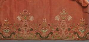  An antique terracotta red textile featuring symmetrical white, green, and hints of yellow and blue floral embroidery or print, with vase-like motifs and whimsical foliage patterns along the bottom edge, exhibiting a traditional and possibly historical design with no known title or artist.