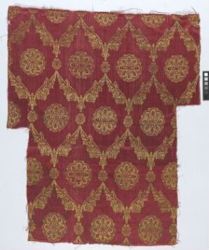  A fragment of antique fabric with a symmetrical pattern of golden medallions connected by diamond-shaped motifs on a deep burgundy background. The design is intricate with radiating golden lines and dots, dominated by warm, luxurious colors, and the edges of the fabric appear frayed and worn.