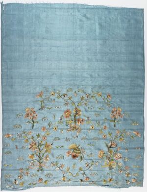  A pale blue piece of textile with a symmetrical floral design in yellow, peach, and green hues towards the lower half, resembling a serene sky-colored background with a spring meadow of flowers. Artist and title unknown.