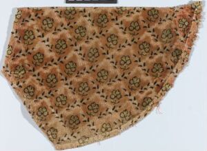  A section of fabric with a curved triangular shape, featuring a dense, floral-patterned design in shades of peachy beige, brown, and goldenrod, suggesting an antique or cultural textile with frayed edges.