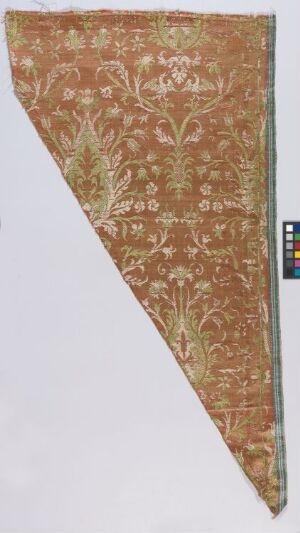  A triangular fragment of aged fabric with an ornate floral pattern in muted reddish-brown, green, and off-white, showing signs of wear and fraying, with a color calibration scale on the right side. Artistname and title are unknown.