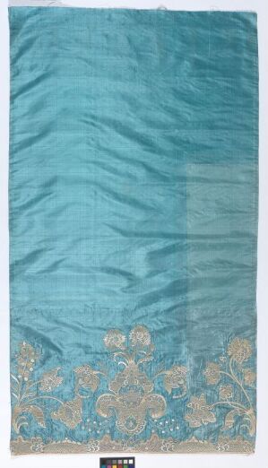  An image of a wrinkled, light teal fabric with a decorative floral border in pale gold, light brown, and off-white colors at the bottom, possibly a textile art piece or a furnishing fabric. The fabric is displayed vertically.